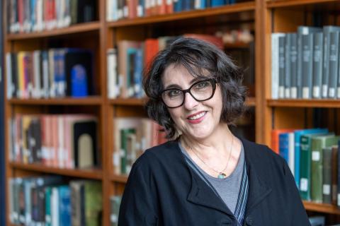 Kristin Sánchez Carter wearing a black jacket over a gray shirt with large bookcases in the background.