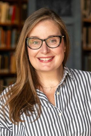 Una Miller wearing a gray and white striped button down collared shirt and black rimmed eyeglasses with a bookcase in the background.