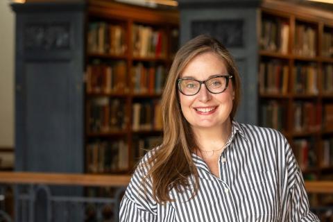 Una Miller wearing a gray and white striped button down collared shirt and black rimmed eyeglasses with a bookcase in the background.