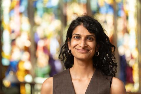 Portrait of a person with long black hair and olive brown blouse smiling with a blurry stained glass window in the background.