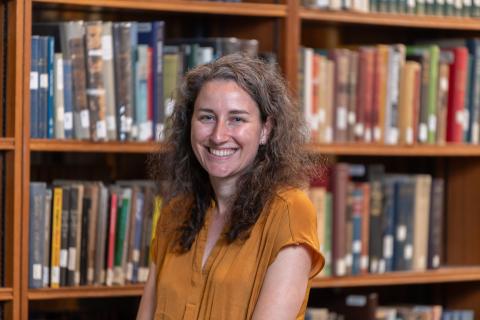 Person with long curly brown hair standing in front of a book case.