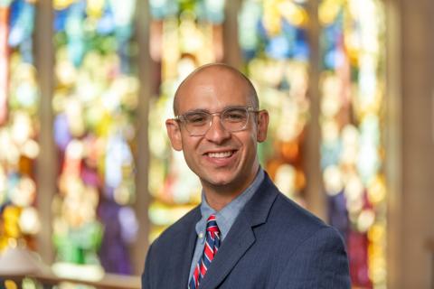 Portrait of a person in a blue suit and glasses in front of a stained glass window.