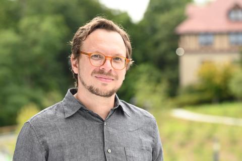 Portrait of a person outside with trees in the background wearing a grey, button-down, collared shirt with glasses and a thin mustache and beard.