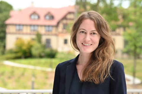 A portrait of a person with long brown/blonde hair with a dark suit jacket standing outside with a slighted blurred building and trees in the background. 