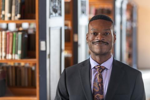 A person standing in a library with book shelves in the background wearing a suit and tie. The person has short cropped black hair and a tight black mustache.