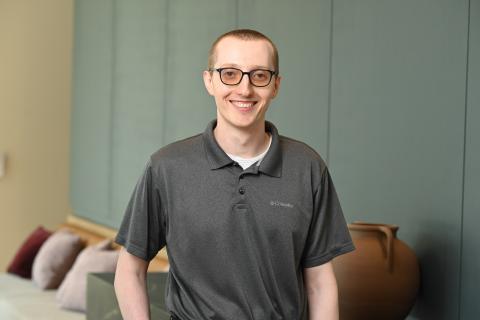 Person wearing a gray collared shirt over a white shirt, black framed eyeglasses, and short light hair. There is part of a couch with pillows and a large clay vase on a table against a green wall. 