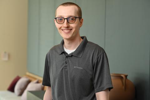 Person wearing a gray collared shirt over a white shirt, black framed eyeglasses, and short light hair. There is part of a couch with pillows and a large clay vase on a table against a green wall. 