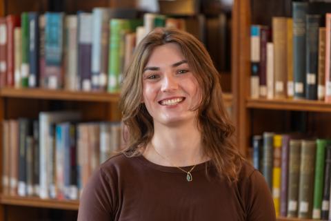 Person with long brown hair, smiling wearing a brown shirt and necklace with a bookshelf in the background.