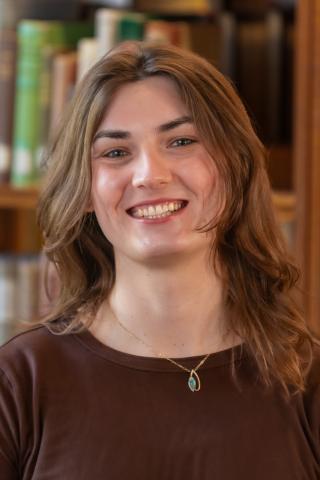 Person with long brown hair, smiling wearing a brown shirt and necklace with a bookshelf in the background.