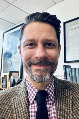 Matthew Schultz with a beard and mustache, wearing a purple and white checked shirt, tan patterned jacket, and dark blue tie with a bookcase and framed art in the background.