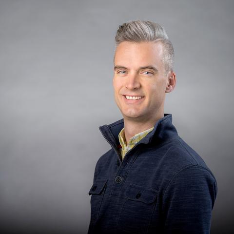 Colin Echeverría Aitken wearing navy shirt with high collar against a gray background.