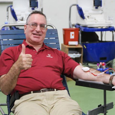 head fencing coach Bruce Gillman giving a thumbs up sign as he sits in a chair donating blood.