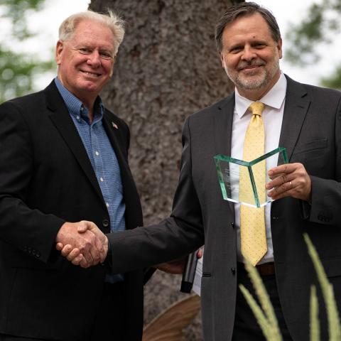 Two men standing in front of a tree shaking hands, one holding a clear glass award shaped like a book