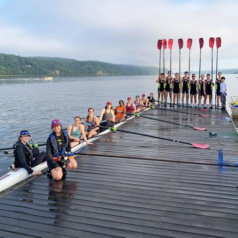 A dock on a lake with hills in the background. In the water a rowing scull is filled with people and five people on the dock holding a paddle.