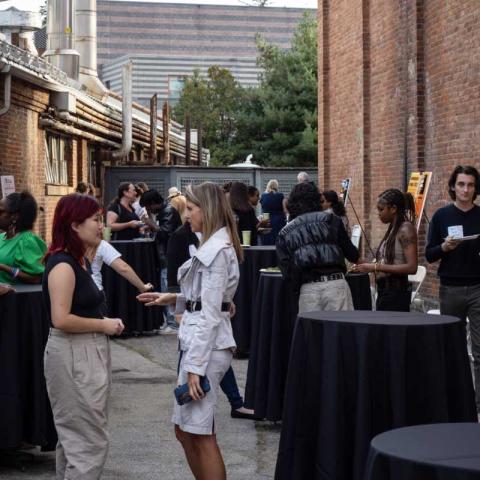 A gathering of people in an open space framed by brick industrial buildings. This is an event with small black cafe tables. People stand around the tables talking.