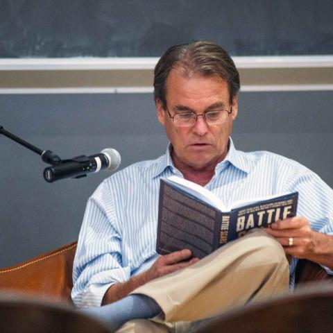 A person with glasses seated behind a microphone reading from a book with the work, Battle, and the cover.