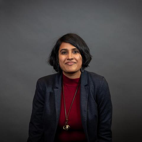 A person with medium length dark hair, dark skin, smiling, wearing a dark blue blazer, red mock-neck shirt, and long necklace with a gold pendant.