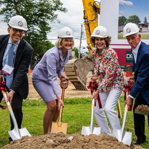 4 people in hard hats smiling and pushing shovels in the ground.