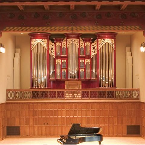 a concert hall with a grand piano on a wooden stage. Behind the stage is a large, ornate pipe organ with polished metal pipes and intricate woodwork.