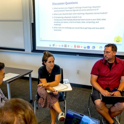 People seated in a circle, some with laptops, in front of a presentation screen, having a discussion.