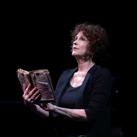Person standing on a dark stage holding an old book and reading out loud.