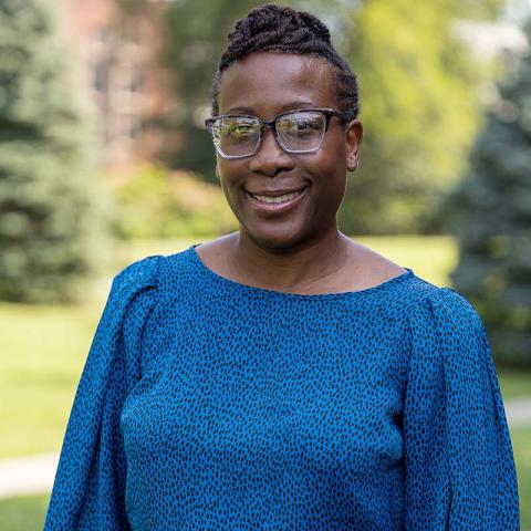 A person with short black hair and glasses stands outside smiling at the viewer. The person is wearing a blue shirt.