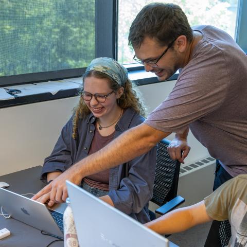 A smiling professor points at a laptop screen while two students look on.