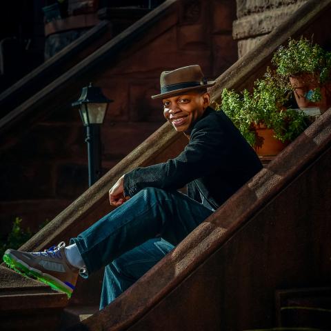 Robert Jones sitting on cement stairs wearing a green shirt, jeans and a light brown hat with a couple of green plants on the stairs behind him.