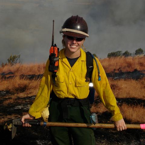 Pictured: Samantha Orient ’05. A person standing with fire-fighting gear (caution-yellow shirt and overalls) holding hatchet/shovel.