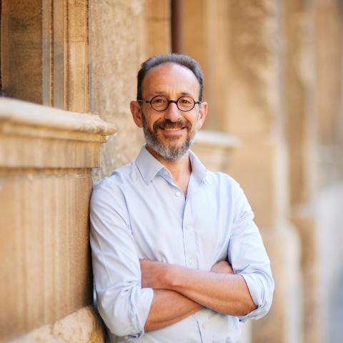 Man leaning up against a stone building wearing a light colored button up shirt and eyeglasses. 