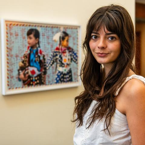 Julia Pippenger with long dark hair and wearing a white strappy shirt, standing next to Indigenous artwork of two young people. 