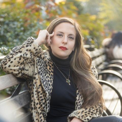 A person sitting on a bench wearing a black shirt, a leopard print jacket with an arm on top of the bench and hand on head. Trees are in the background. 