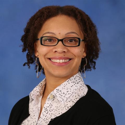 Jennifer Sims wearing a white collared shirt with dark spots, a black sweater, black rimmed eyeglasses and large shaped earrings against a blue background.