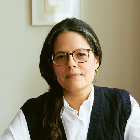 Person with long dark hair and classes sitting at a table with their arms crossed.