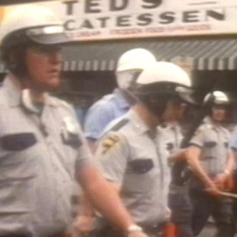 A blurry frame from the film "Power" by Yance Ford. It shows a group of police officers in helmets, holding batons and marching in a line.