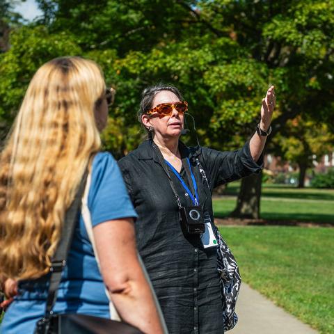 Person with rose colored glasses standing outside in the Vassar Quad speaking with their hand raise while giving a tour with others looking on.