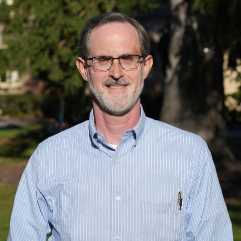 Ken Foster wearing a light blue with white striped collared shirt, eyeglasses, and short hair with beard and mustache with a brick building and large tree in the background.