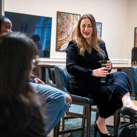 Dr. Michelle Monje ’98, a person with long blond hair and a dark dress, sits in a room talking to some other people.