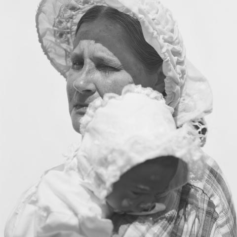 A close-up photograph of a woman holding a small child. They wear historical clothing.