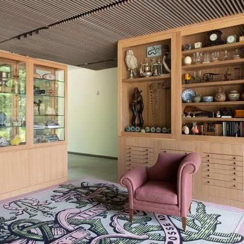 A staged room with a cusioned chair and rug with two wood and glass cabinets filled with random, mixed items flanking the rug and chair.