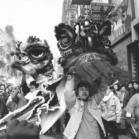 A black and white photo of a bustling city street during a parade with a Chinese Dragon being carried.