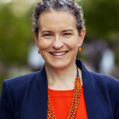 Person wearing a orange shirt, blue jacket, orange beaded necklace, round orange earrings, and has short hair. 