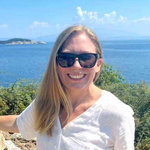 A person with long blonde hair, sun glasses, and a white shirt smiling with water and an island in the background.