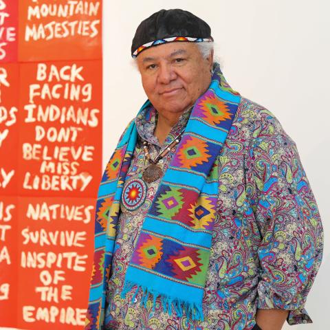 Photo of artist Edgar Heap of Birds standing in front of one of his text-based works. 