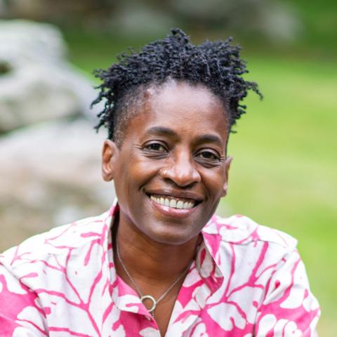 Person with dreadlocks and bright pink flowered shirt sitting outside on a low rock wall.