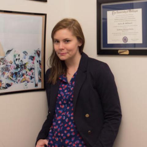 Dr. Andrea McDonnell, a person with long brown hair and a black coat, stands in front of a wall with diplomas and a painting hung on it.