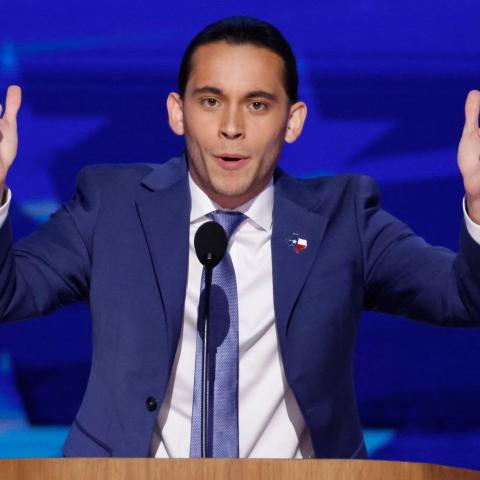 Person standing behind and lectern and microphone with both arms in the air speaking emphatically.