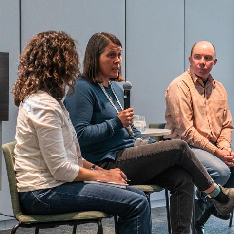 Panel of three people sitting on a stage with one person speaking into a microphone while the others look on.