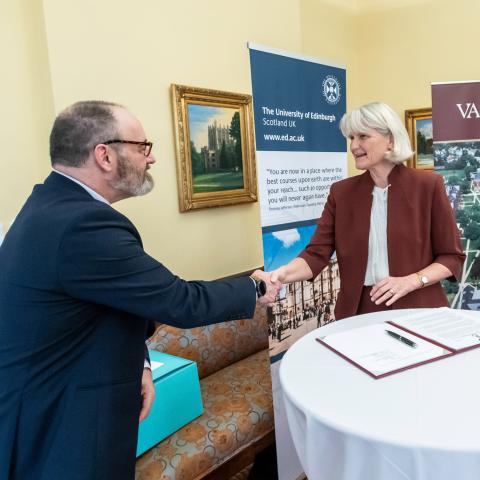 Two people shaking hands over a table with two banners behind them that read: University of Edinburgh and Vassar.