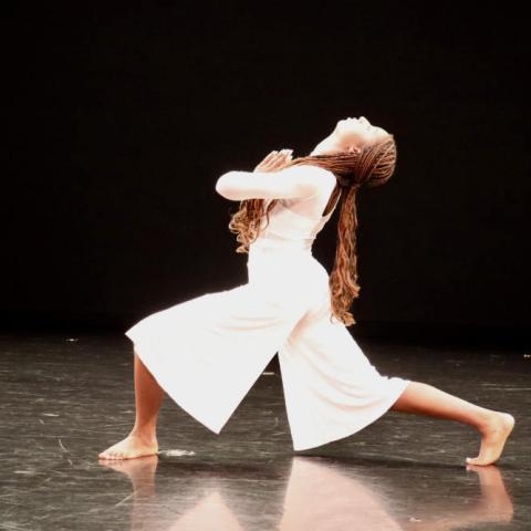 A person in a white dress and long dark hair performs on a dark stage.